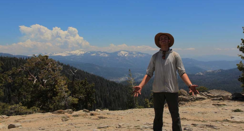 A person stands on a rocky overlook. Behind them are the snow capped mountains of the high sierra. 